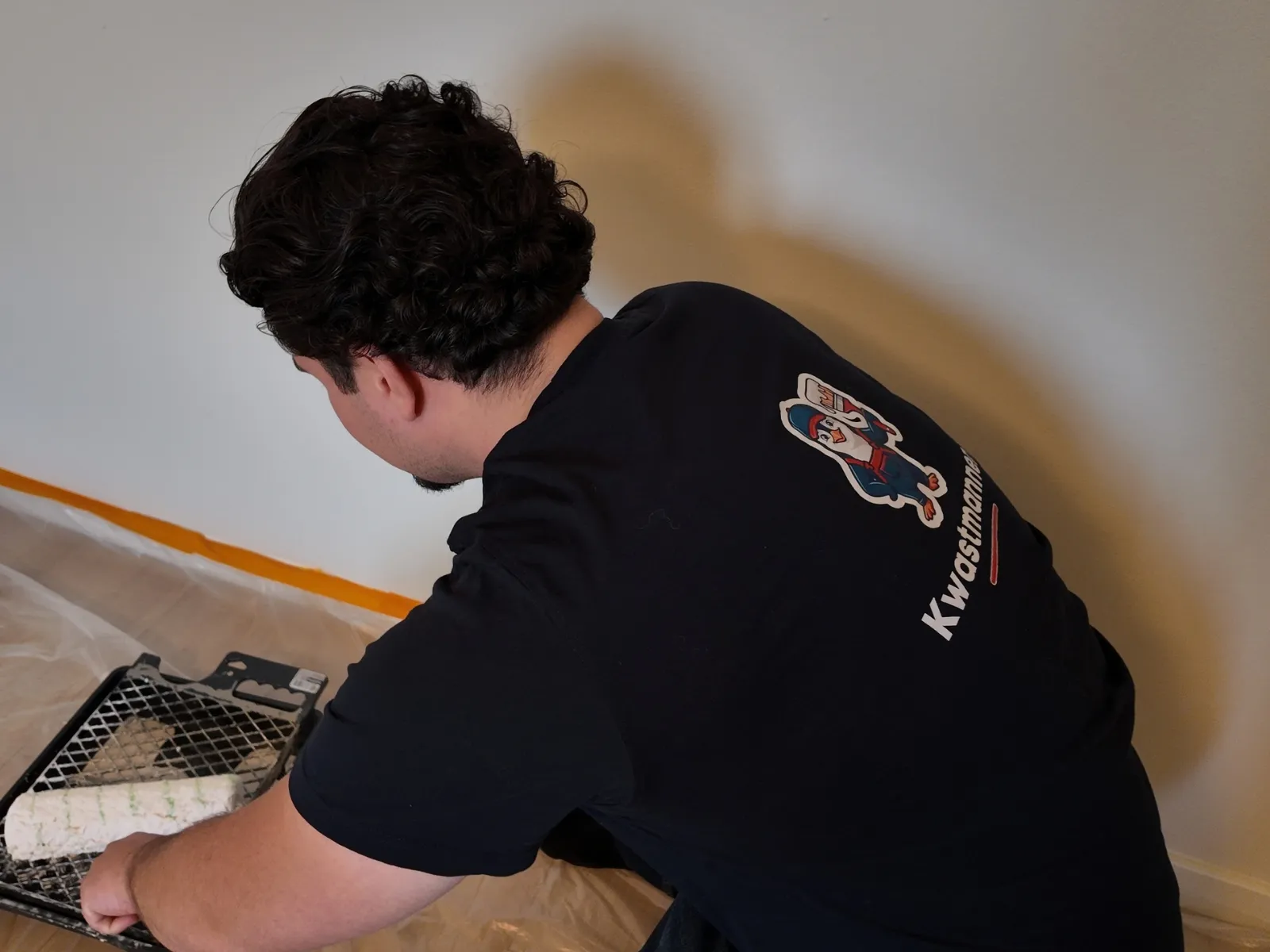 Schilder van Kwastmannen buigt zich over de verfbak om de roller te vullen, de vloer afgedekt met folie en de plint afgeplakt met tape.