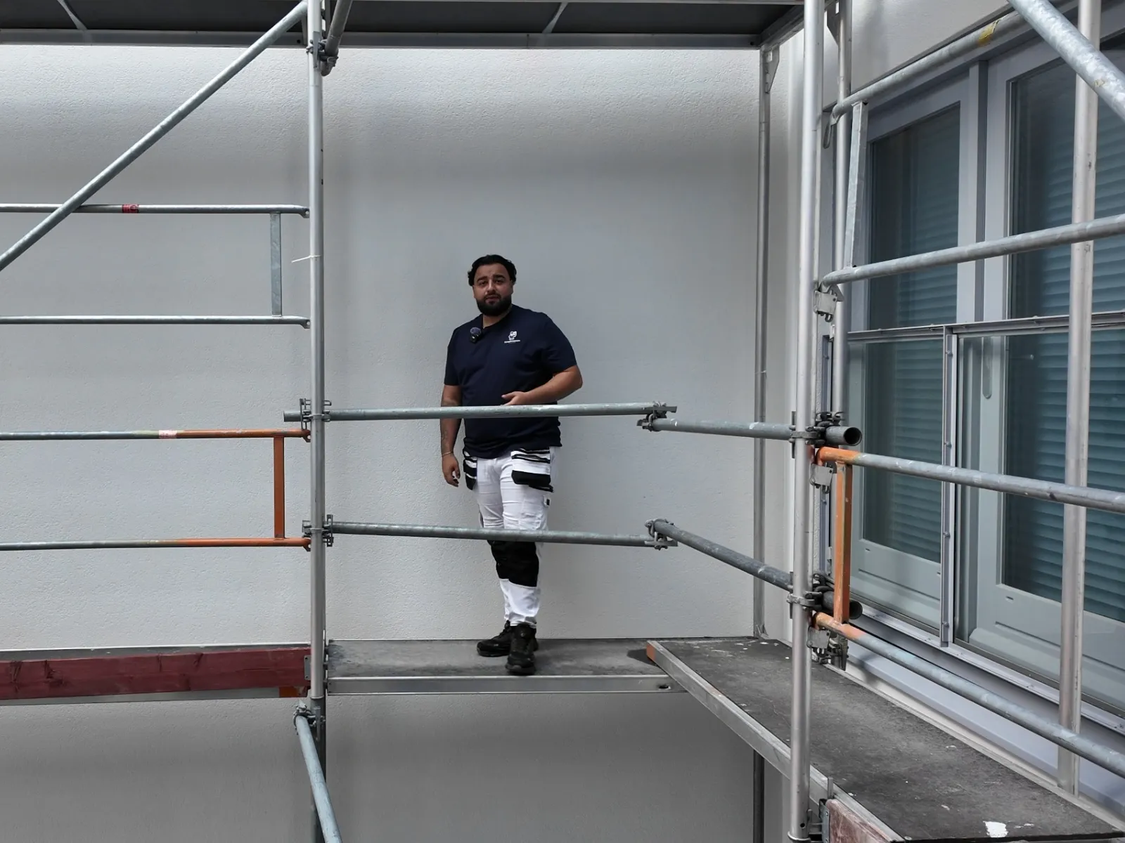 Schilder in werkkleding op de steiger voor een pas afgewerkte gevel, met het kozijn en de balkondeur naast hem al geschilderd.