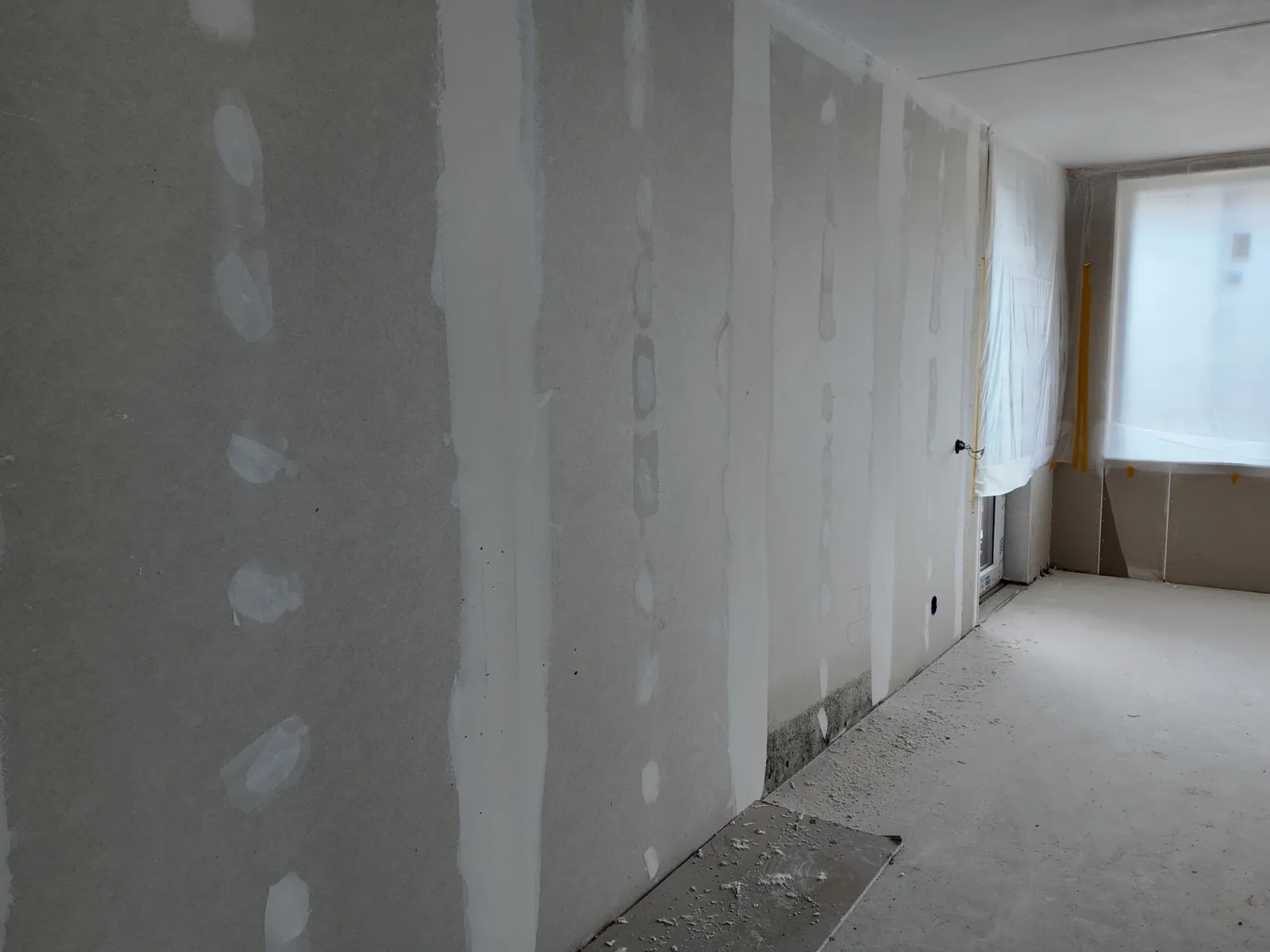 Lege kamer voor het schilderen: kale gipsplaten met afgeplakte naden en gevulde schroefgaten, het raam afgedekt met folie