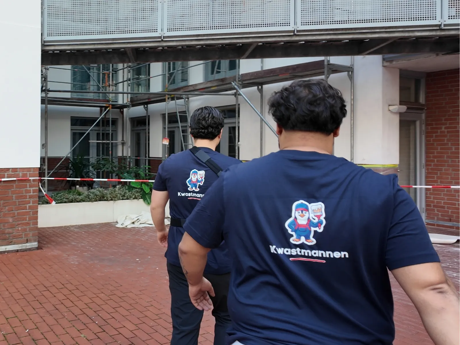 Twee schilders van Kwastmannen lopen in bedrijfsshirt naar het werk, het logo met de pinguïn duidelijk op de rug, op de achtergrond een pand in de steigers