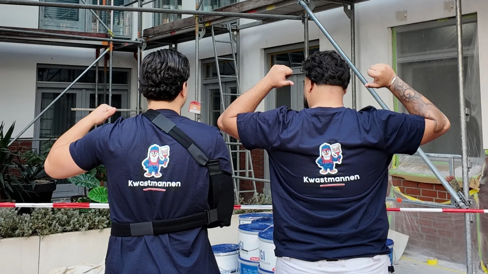 Twee schilders van Kwastmannen met het logo op de rug bij een pand in de steigers, ontspannen poserend met de armen omhoog